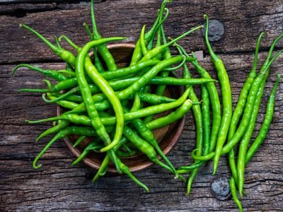 Fresh organic green chili peppers placed in a wooden bowl with some scattered around on rustic wooden surface.
